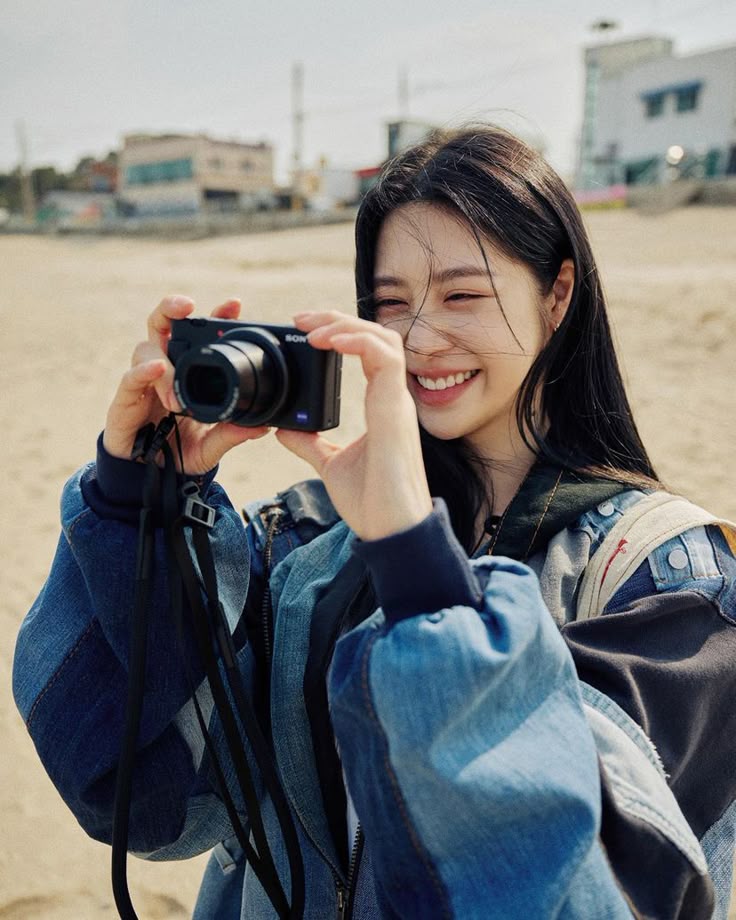 A woman smiling on the beach in a denim jacket, holding a camera under bright sunlight
