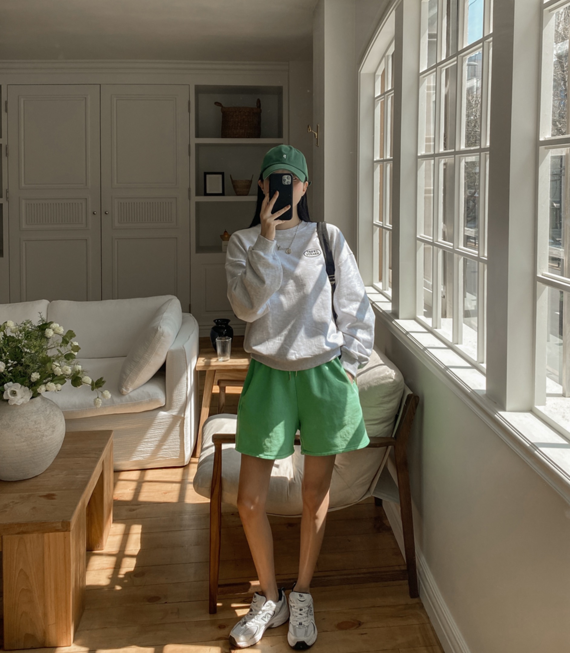 Woman wearing grey sweatshirt, green jogger shorts, and a cap.