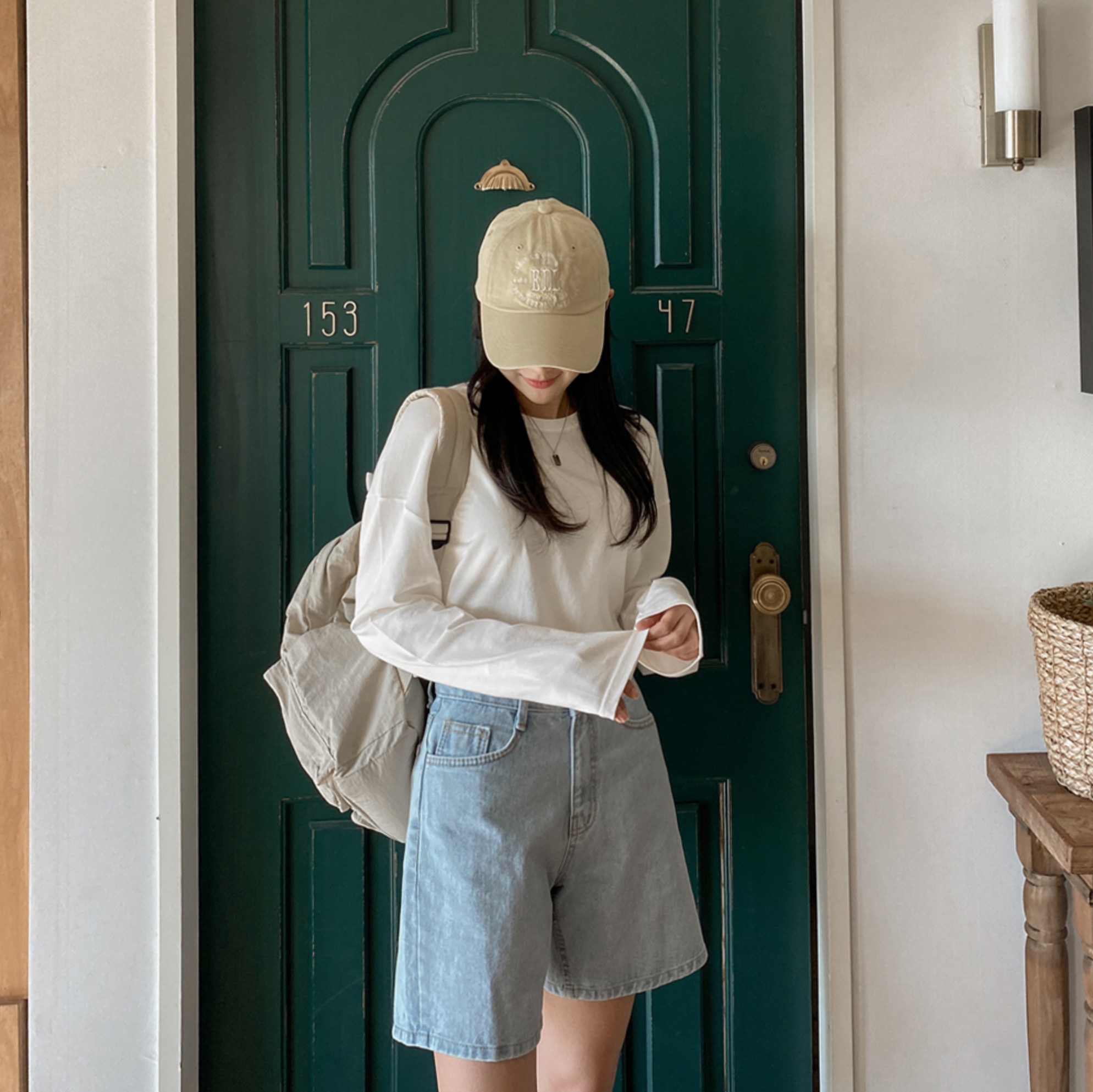 Woman wearing cream long sleeve, light denim shorts, beige cap, and backpack.
